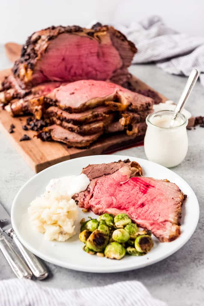 white plate with slice of prime rib, brussels sprouts and mashed potatoes, prime rib roast behind with horseradish sauce to the side