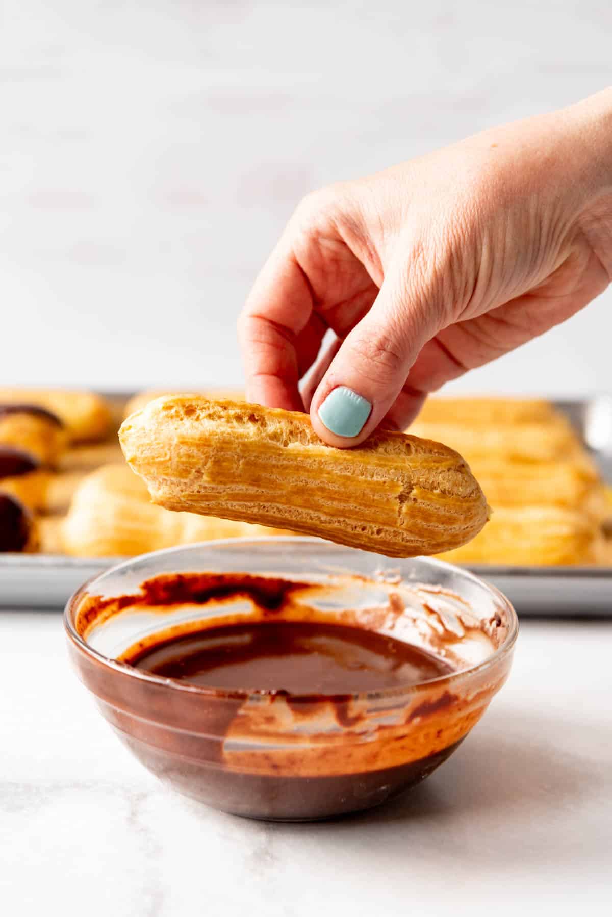 A hand holding a filled eclair over a bowl of chocolate ganache.