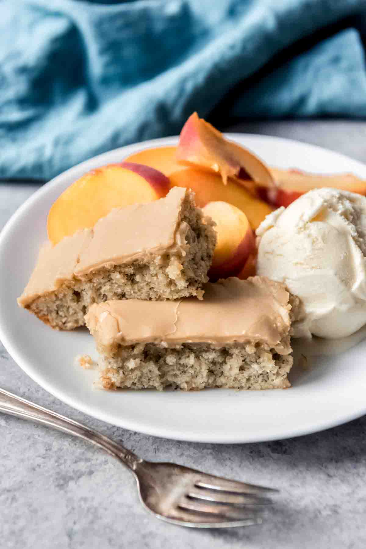 Banana bars with penuche frosting are stacked on a plate with a scoop of ice cream and sliced fruit. 