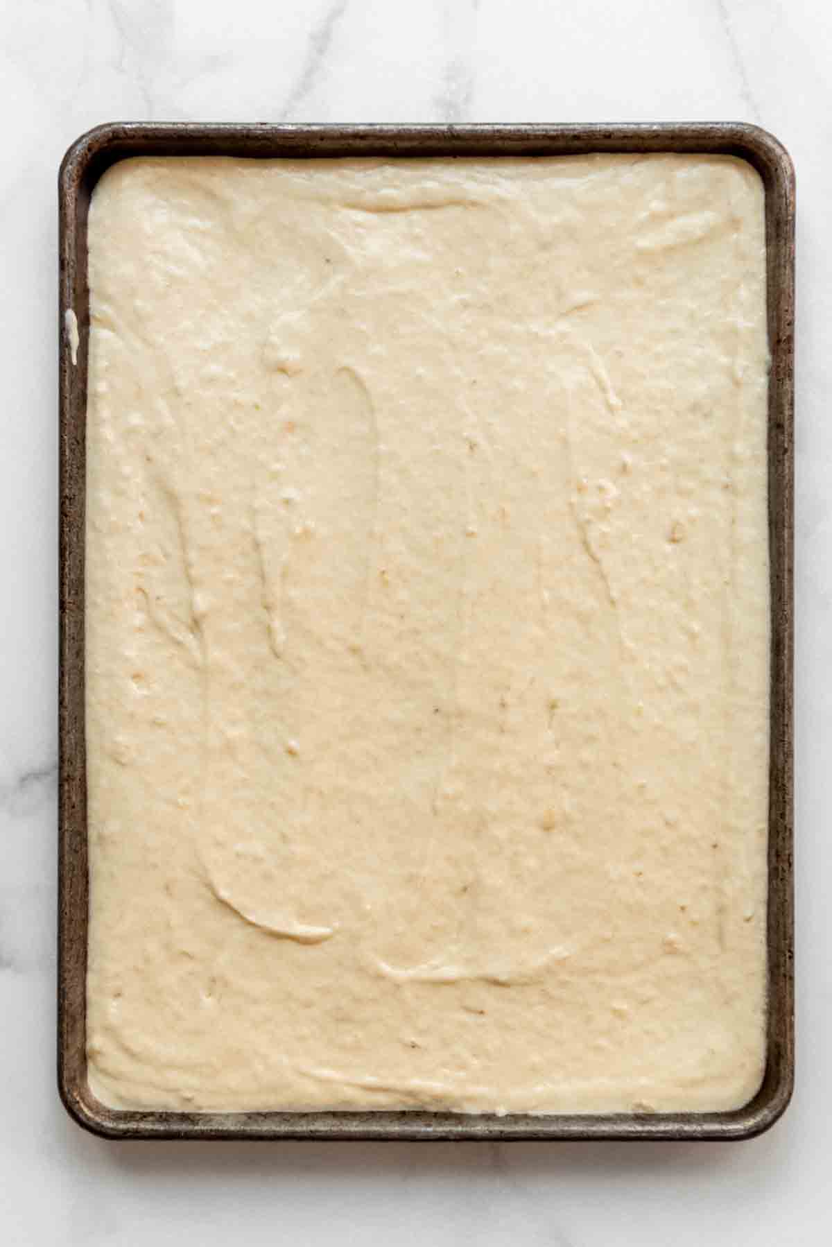 Banana bar batter is spread into a greased half sheet pan, ready to be baked. 