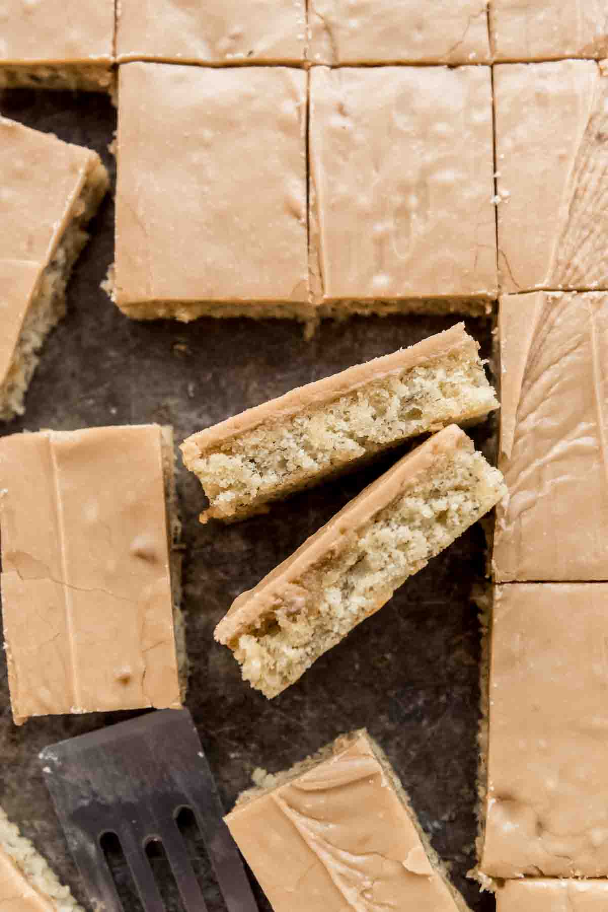 Cut banana bars with penuche frosting are displayed in a metal pan. Two bars are tilted sideways, showing the frosted and banana layers, and a metal spatula is visible at the bottom of the image. 