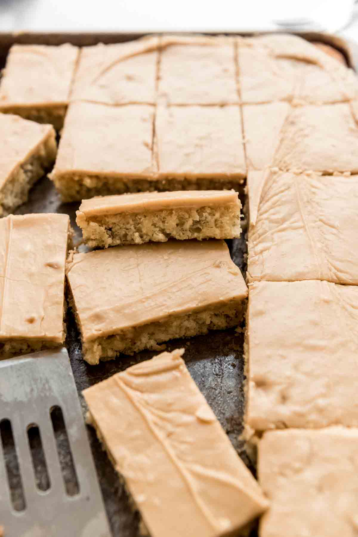 A pan of frosted banana bars are scattered in a metal tray, with a metal spatula visible in the corner. 