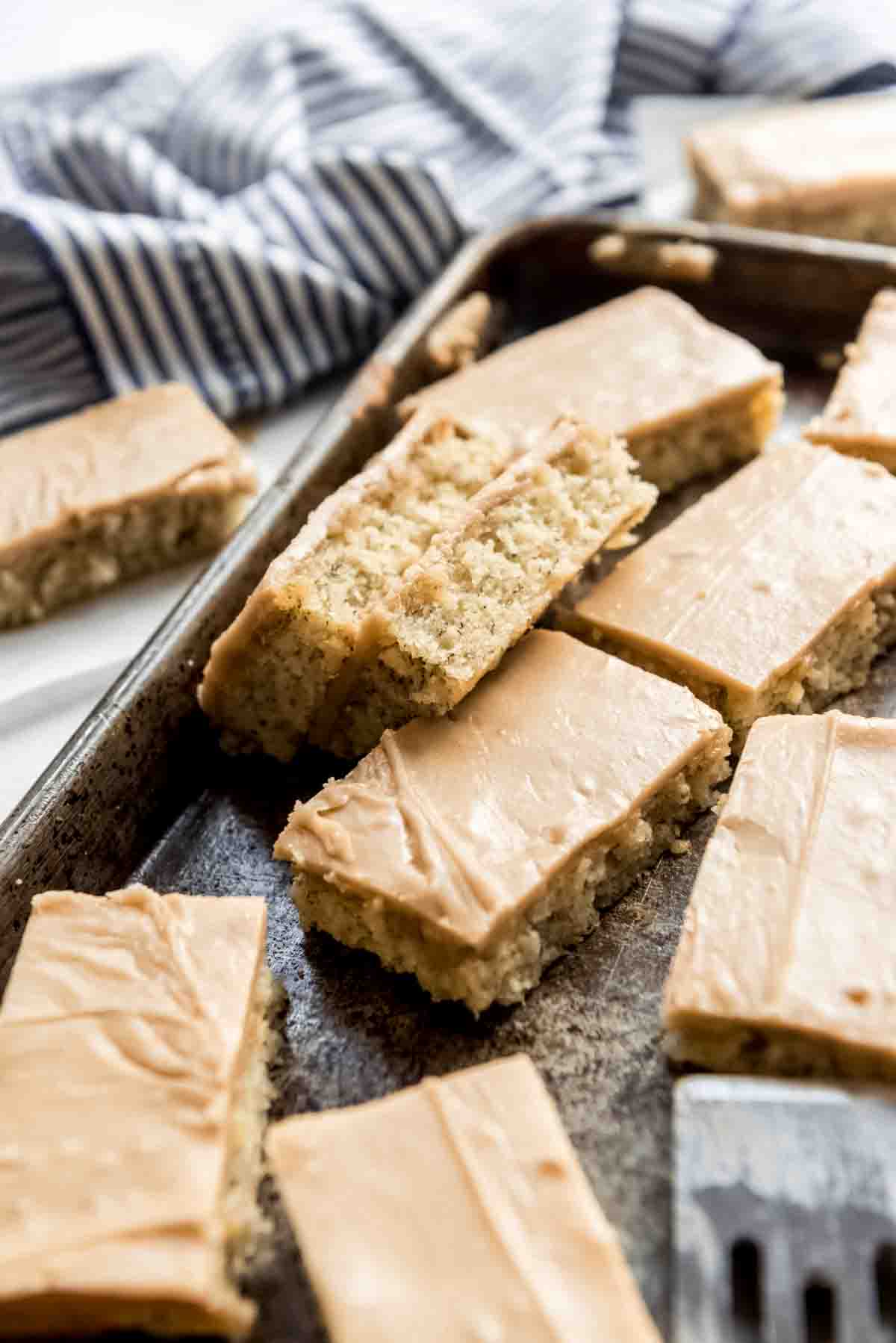 Cut and frosted banana bars are scattered in a metal baking pan. Two bars are turned on their sides, making the bottom banana base visible, the frosted tops are showing on other bars. 