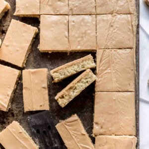 Cut banana bars with penuche frosting are displayed in a metal pan. Two bars are tilted sideways, showing the frosted and banana layers, and a metal spatula is visible at the bottom of the image.