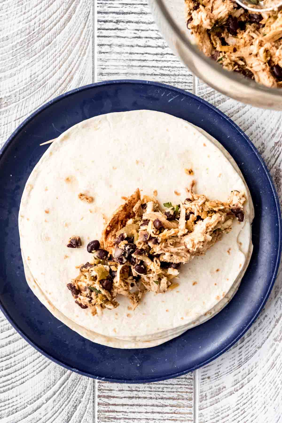 Chicken flautas filling is placed on half of a flour tortilla that is stacked on a blue plate. 