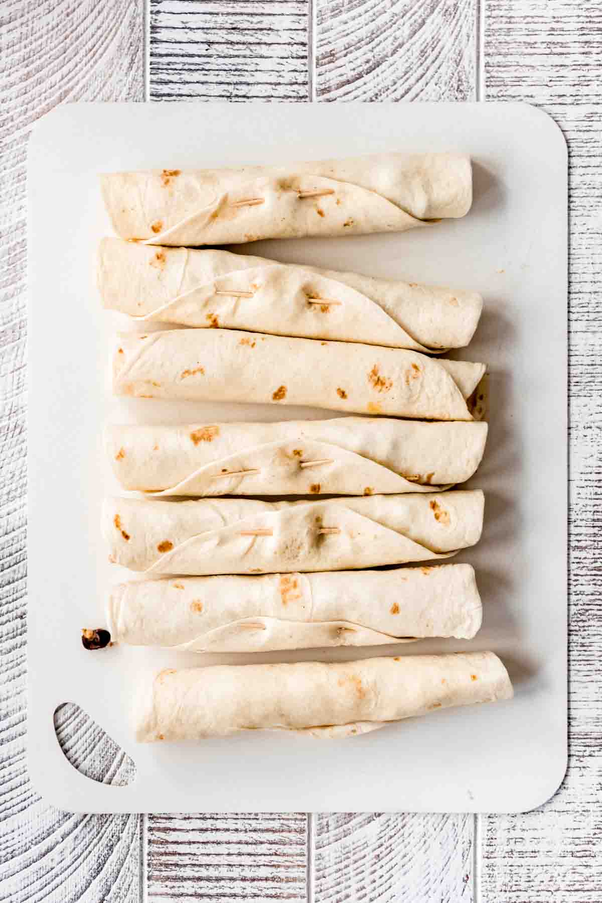 Filled and rolled chicken flautas are lined up on a white cutting board. Toothpicks are visible in some of the rolled flautas. 