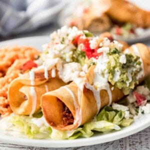 A close up of two chicken flautas that have been fried and topped with sour cream, guacamole, chopped tomatoes and crumbled queso fresco.