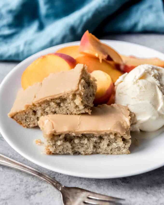 Banana bars with penuche frosting are stacked on a plate with a scoop of ice cream and sliced fruit.