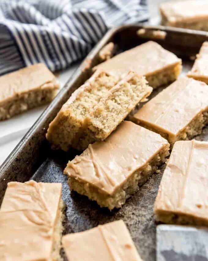 Cut and frosted banana bars are scattered in a metal baking pan. Two bars are turned on their sides, making the bottom banana base visible, the frosted tops are showing on other bars.