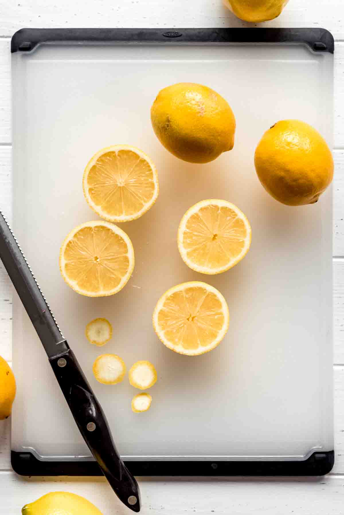 Lemons sliced in half are resting on a cutting board. Two full lemons are ready to be cut, with a serrated knife visible at the bottom of the frame. 