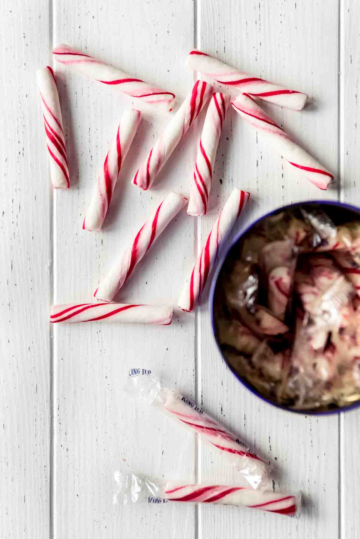 Unwrapped and wrapped peppermint sticks are scattered on a surface, next to a cup containing more sticks. 