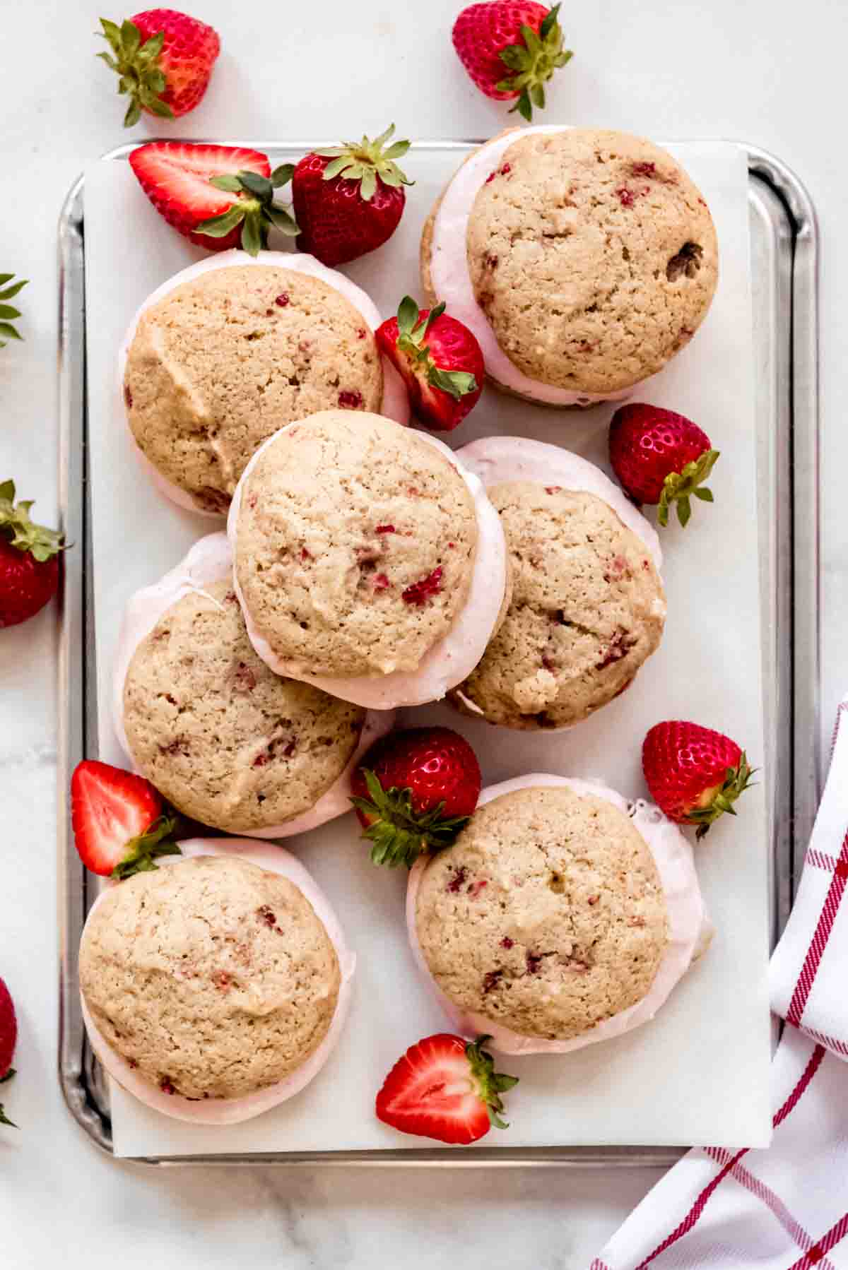 A tray of prepared strawberry whoopie pies are displayed and stacked on each other. Whole and half strawberries are scattered among the whoopie pies. 