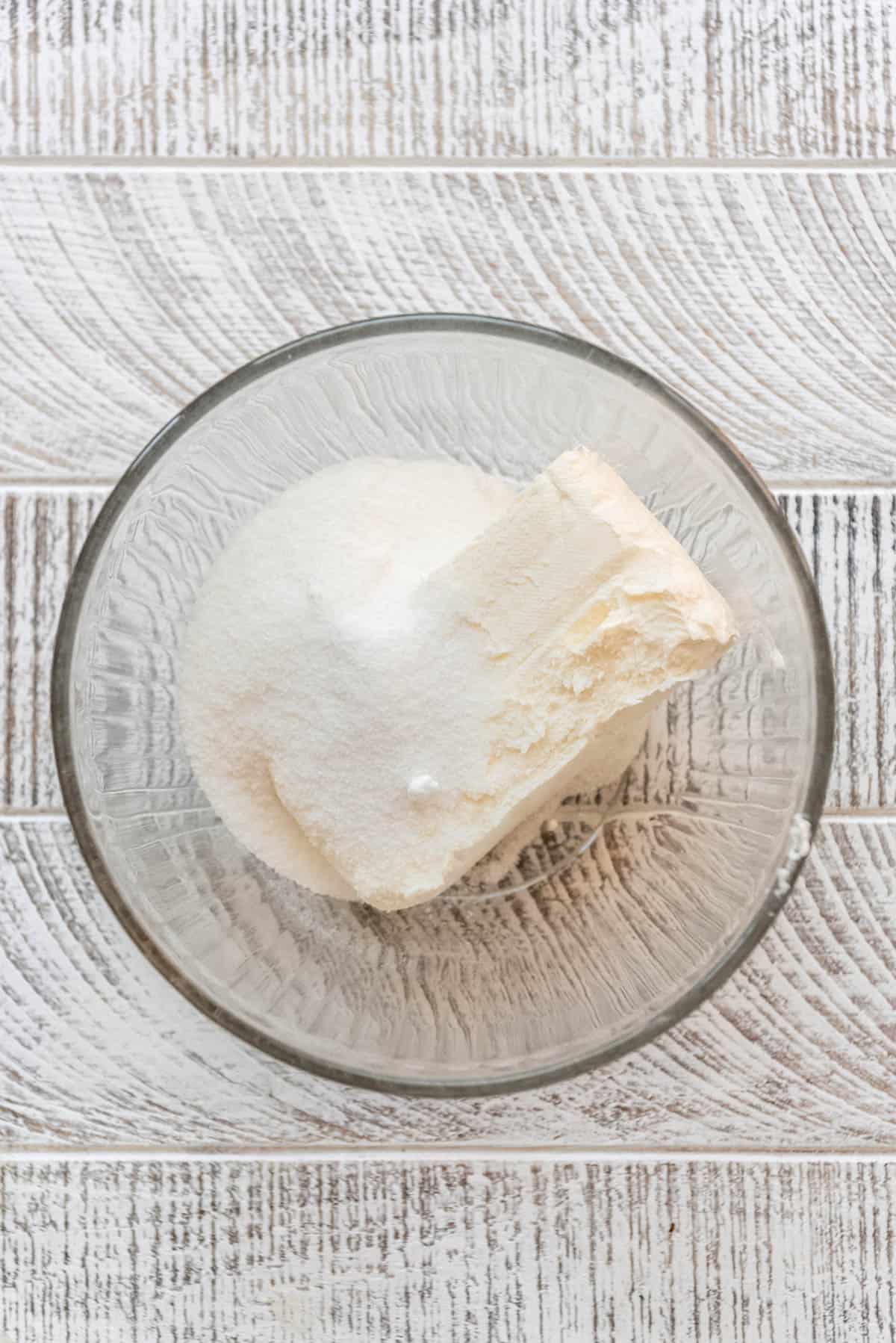 A block of cream cheese and sugar are resting in a clear glass bowl. 