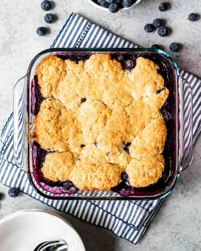 A square baking dish of cooked blueberry cobbler is resting on a striped kitchen towel. Blueberries and serving bowls with spoons are visible around the dish.