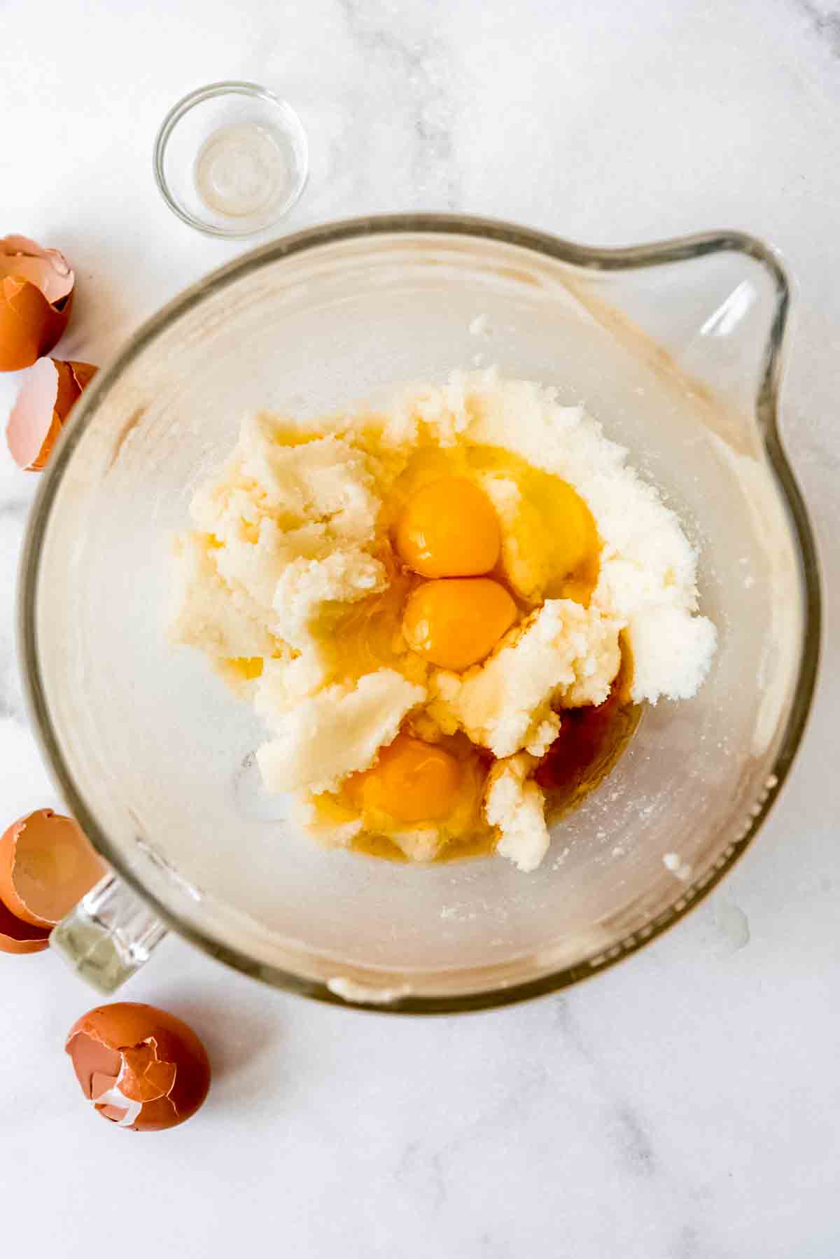 Mixed butter and sugar are in a large glass mixing bowl with eggs and vanilla, ready to be combined together. Cracked egg shells are visible outside of the bowl, with an empty small glass bowl. 