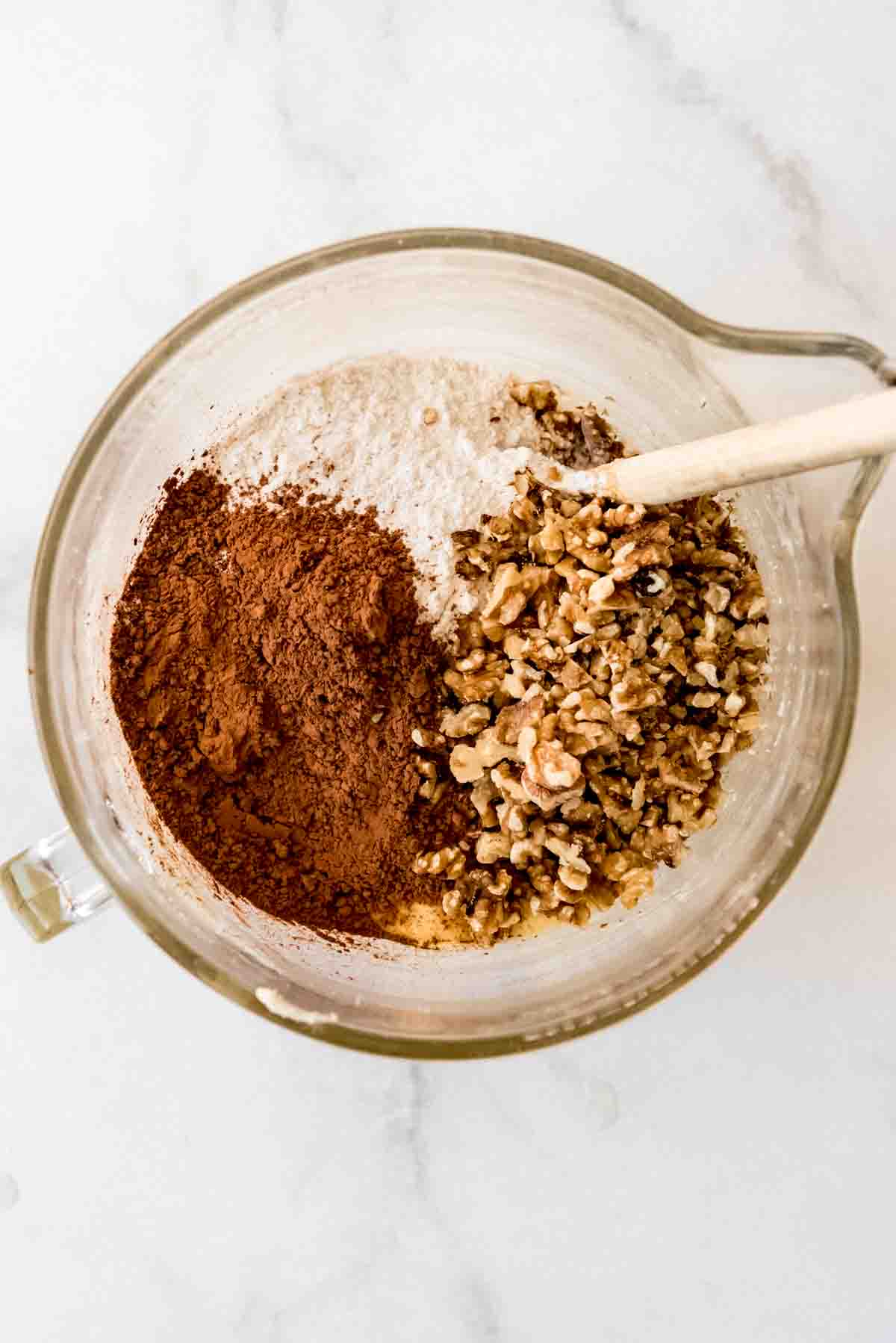 Cocoa powder, flour and chopped walnuts are added to a glass mixing bowl, ready to be blended with a wooden spatula.  