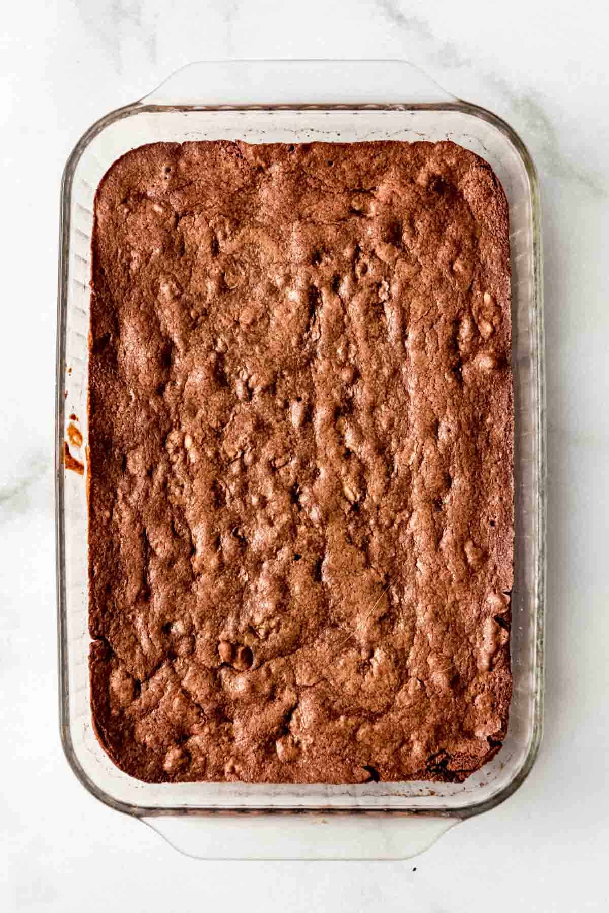 Cooked brownies have been removed from the oven and are resting on a white marbled surface, cooling before frosting is added. 