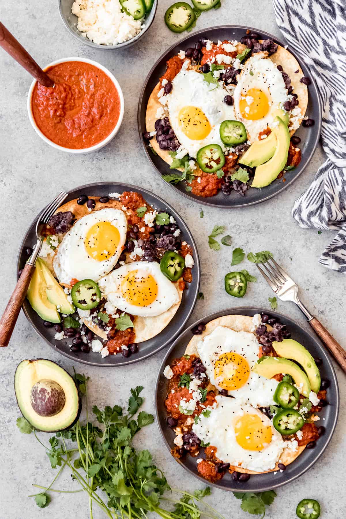 three plates of huevos rancheros served with avocado, jalapeno cheese and cilantro with a side of homemade ranchero sauce in a bowl