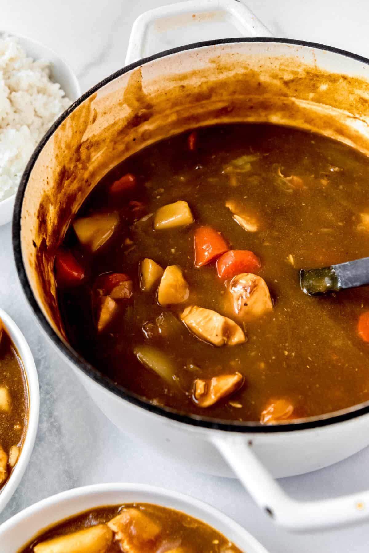 A close up of rich brown curry in a dutch oven. Carrots, potatoes and cubed chicken pieces are visible in the sauce. 