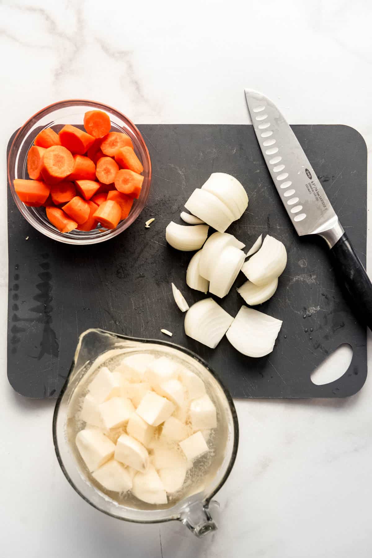 Cubed potatoes, sliced carrots and onion wedges on a black cutting board with a sharp knife on the right edge of the cutting board. 