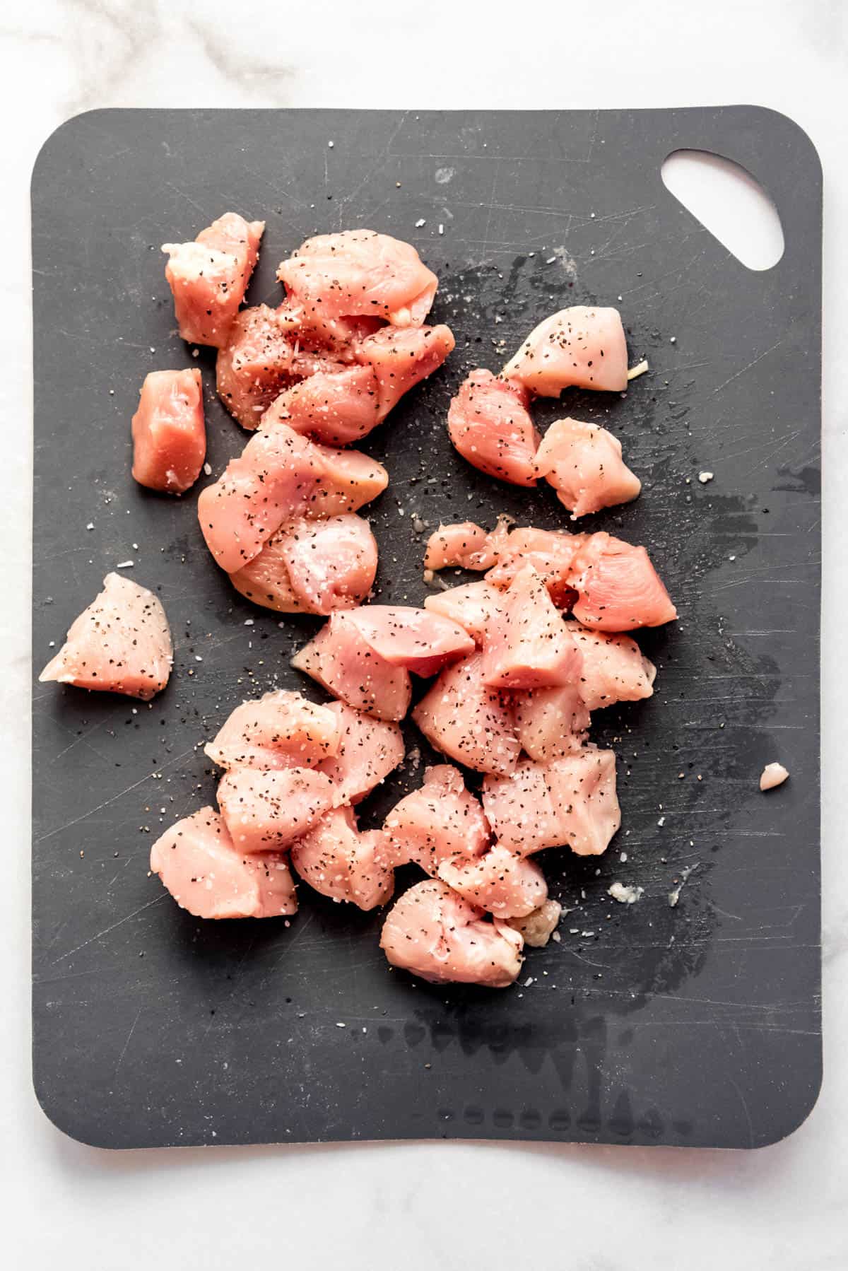 Cubed chicken is resting on a cutting board, after being sprinkled with salt and ground pepper. 