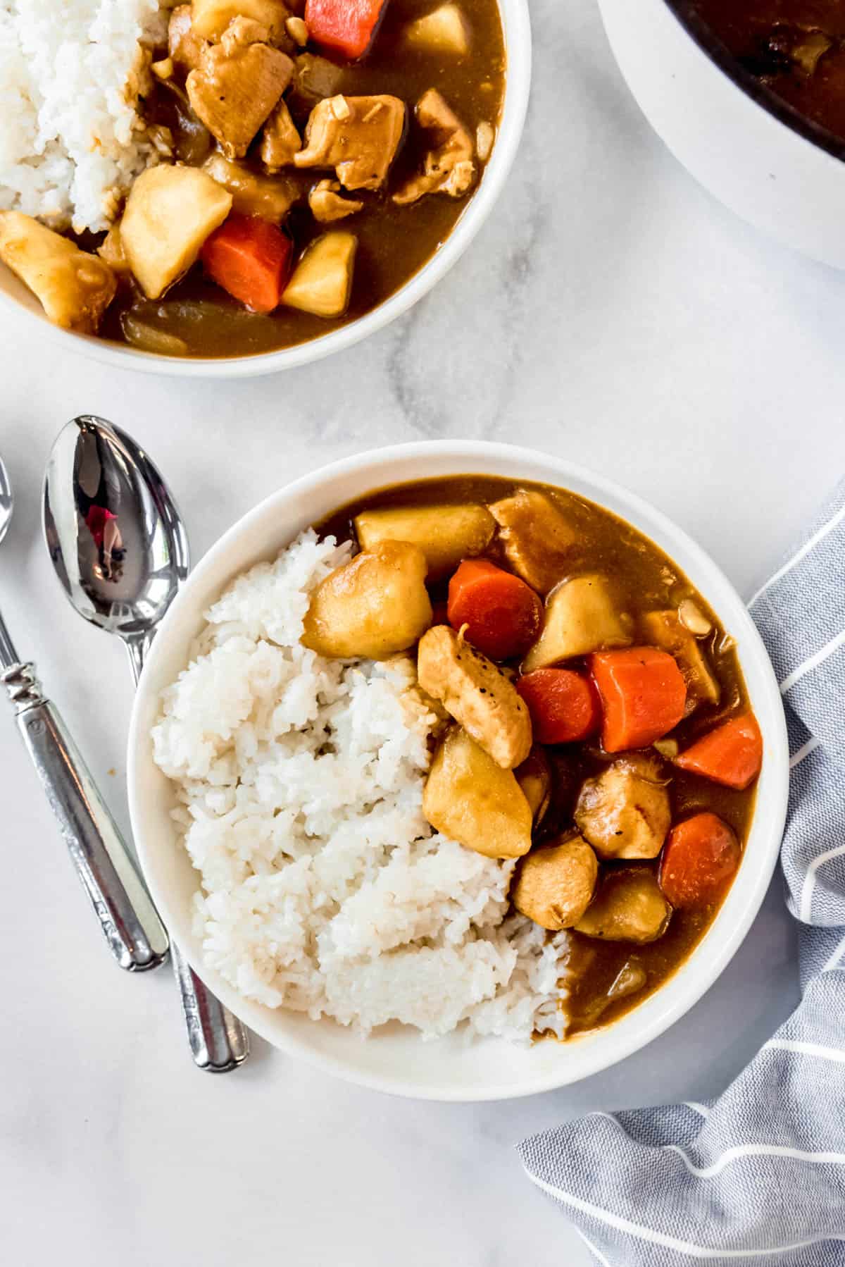 Close up of two bowls of Japanese chicken curry, with cooked chicken, carrots and potato chunks visible, and white rice on the side. 