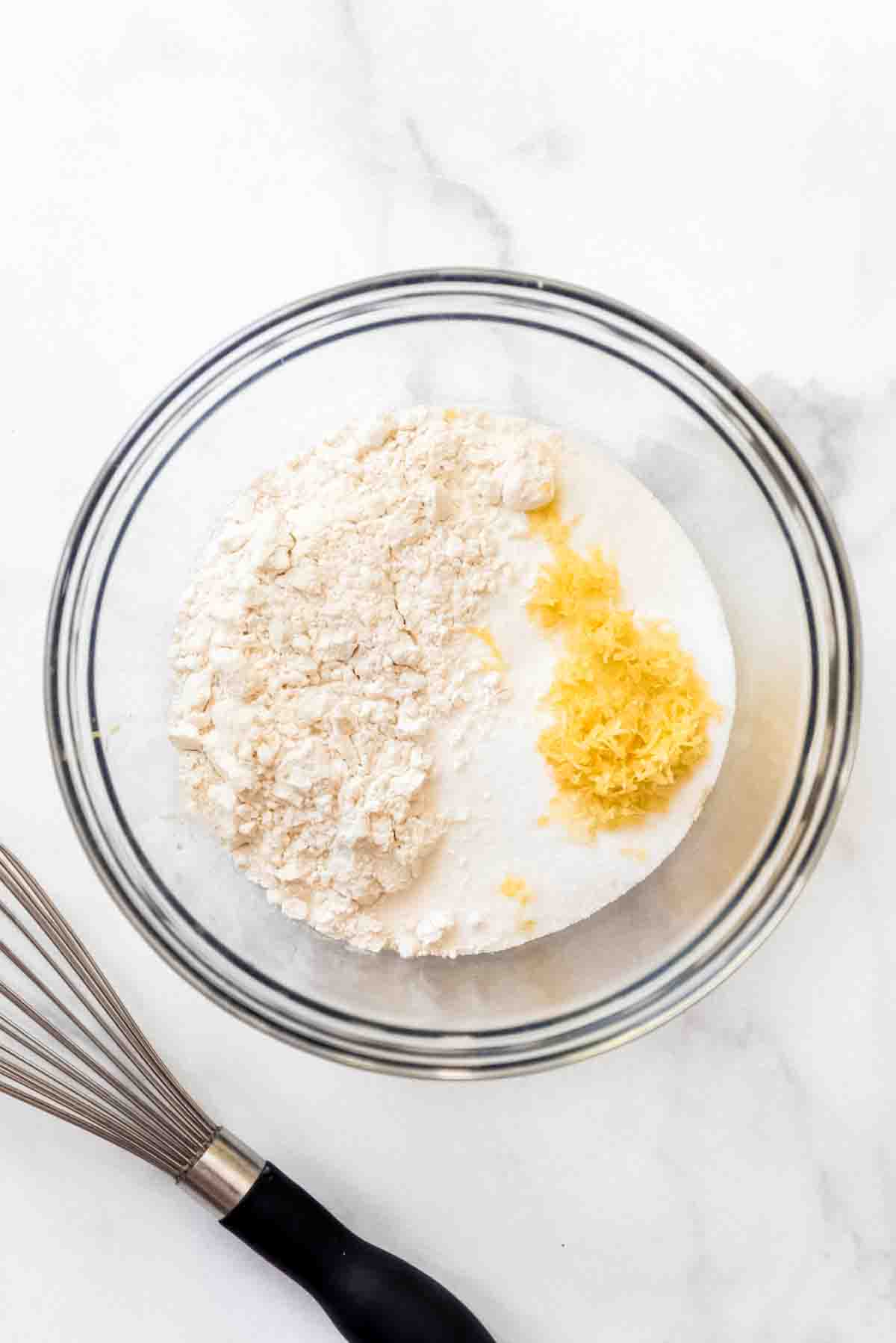 Flour, granulated sugar, and lemon zest are in a bowl, ready to be combined. A metal wisk is visible to the left of the glass bowl, and both are resting on a white marbled surface. 