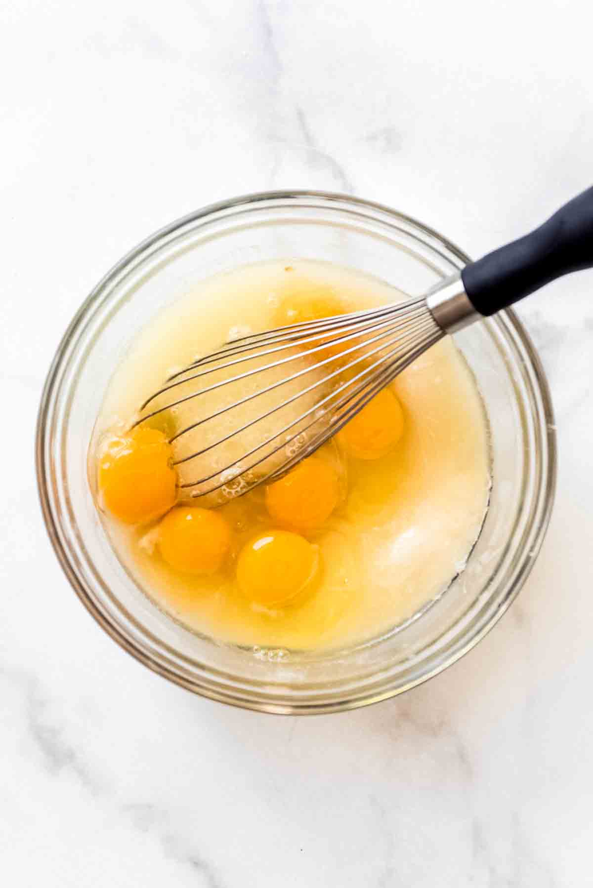 Eggs and lemon juice have been added to a mixture in a clear glass bowl, and a metal whisk is in the bowl, ready to stir the ingredients. 