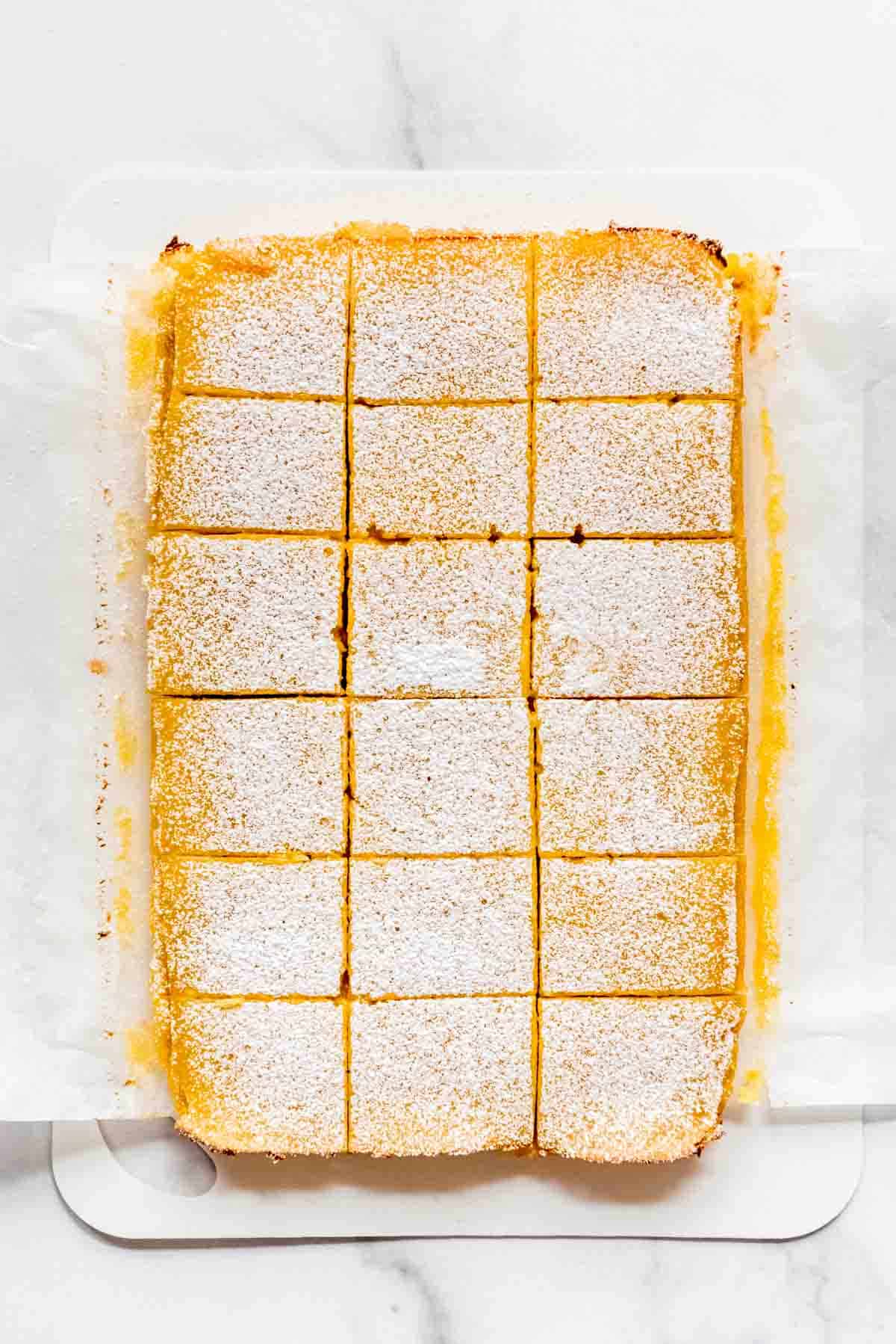 Cooked lemon bars have been removed from the nine by thirteen pan and placed on a white cutting board. The bars have been cut into eighteen squares and are resting on a parchment paper sling. 