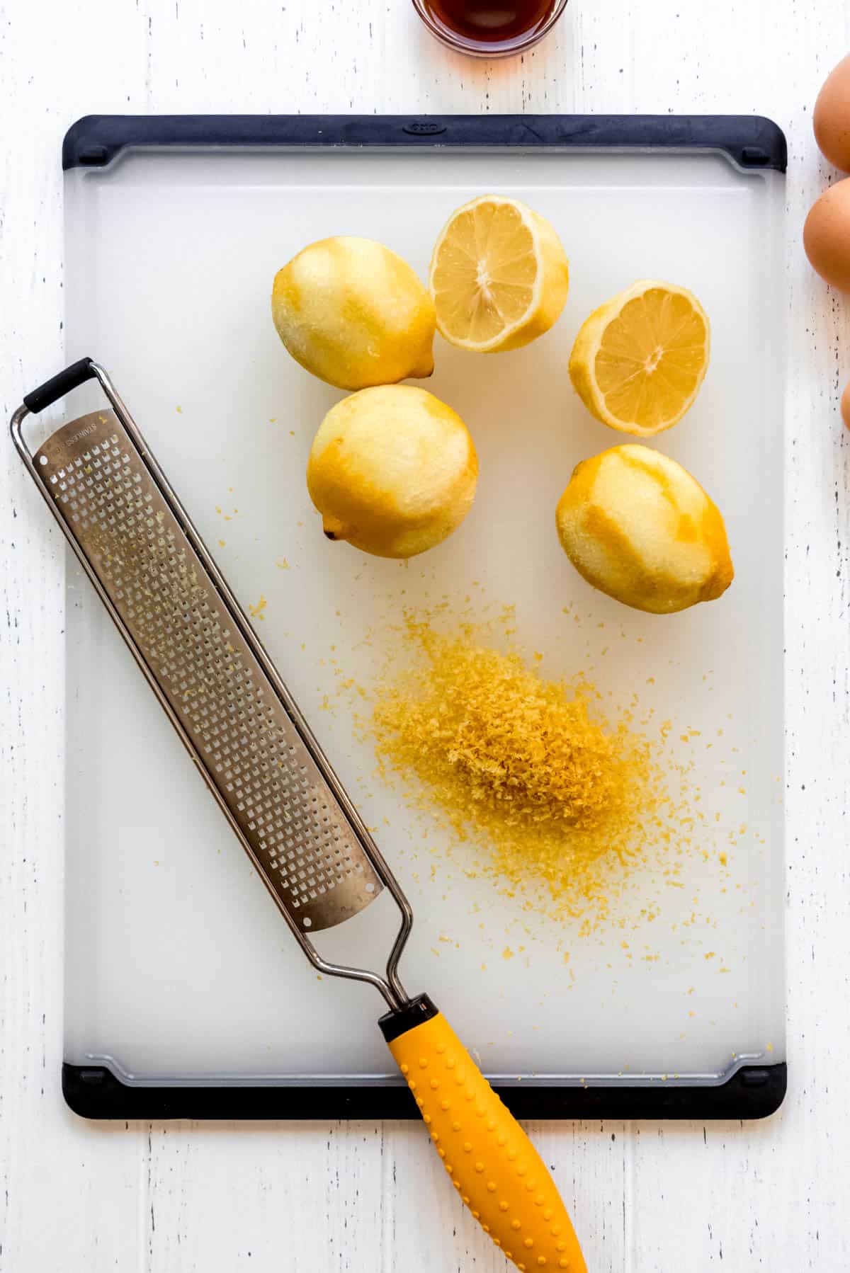 Three whole lemons and one halved lemon are resting on a cutting board next to a microplane. The lemons have been zested, and a pile of lemon zest is on the cutting board. 