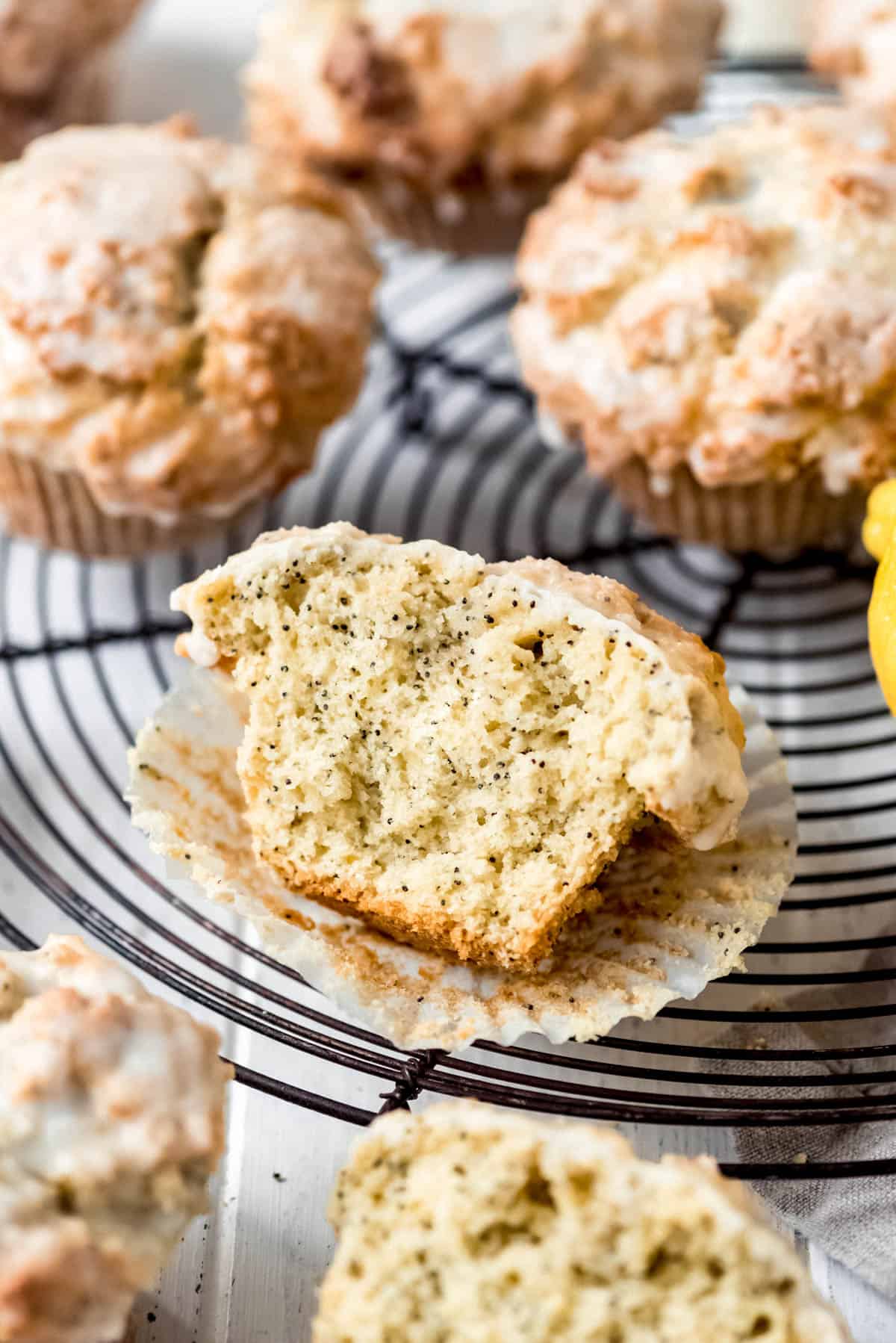 A half muffin is resting on a circular wire rack, displaying poppy seeds throughout the muffin and a glaze on top. Other whole muffins surround it on and off the rack. 