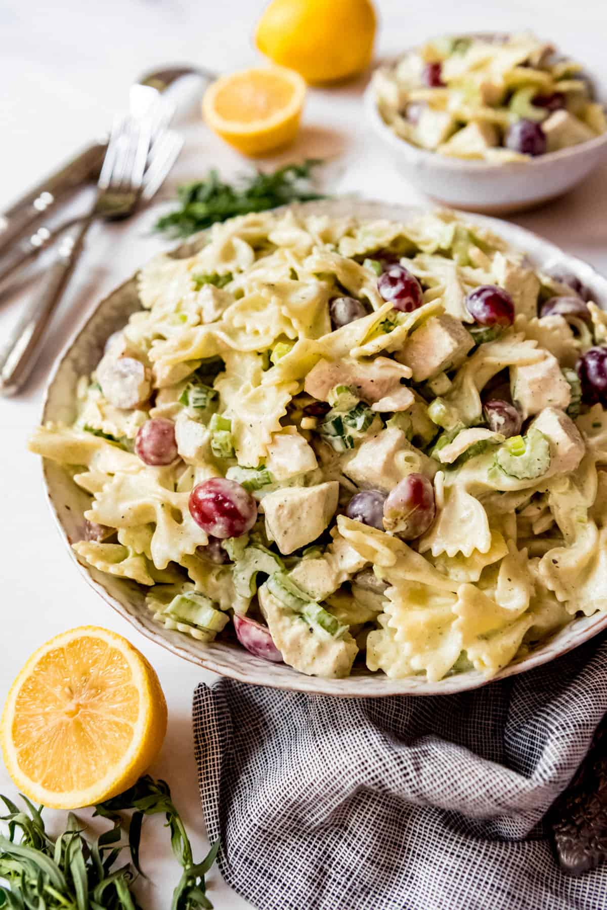 Side shot of Lemon Tarragon Pasta Salad, in stoneware bowl next to lemon and tarragon, with smaller bowl in background