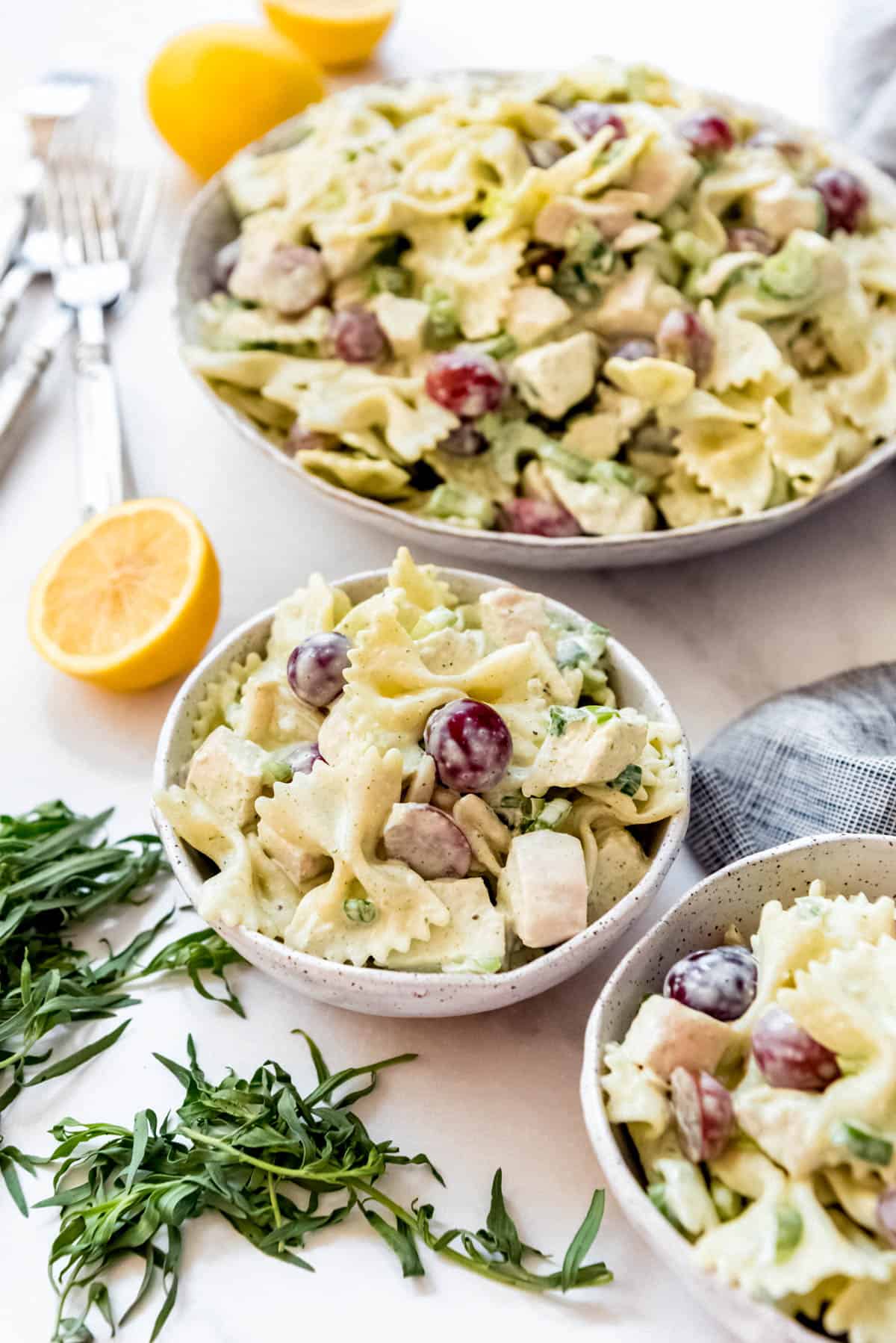 Side shot of Lemon Tarragon Pasta Salad, one large bowl and 2 smaller ones, next to forks, lemons, and tarragon.