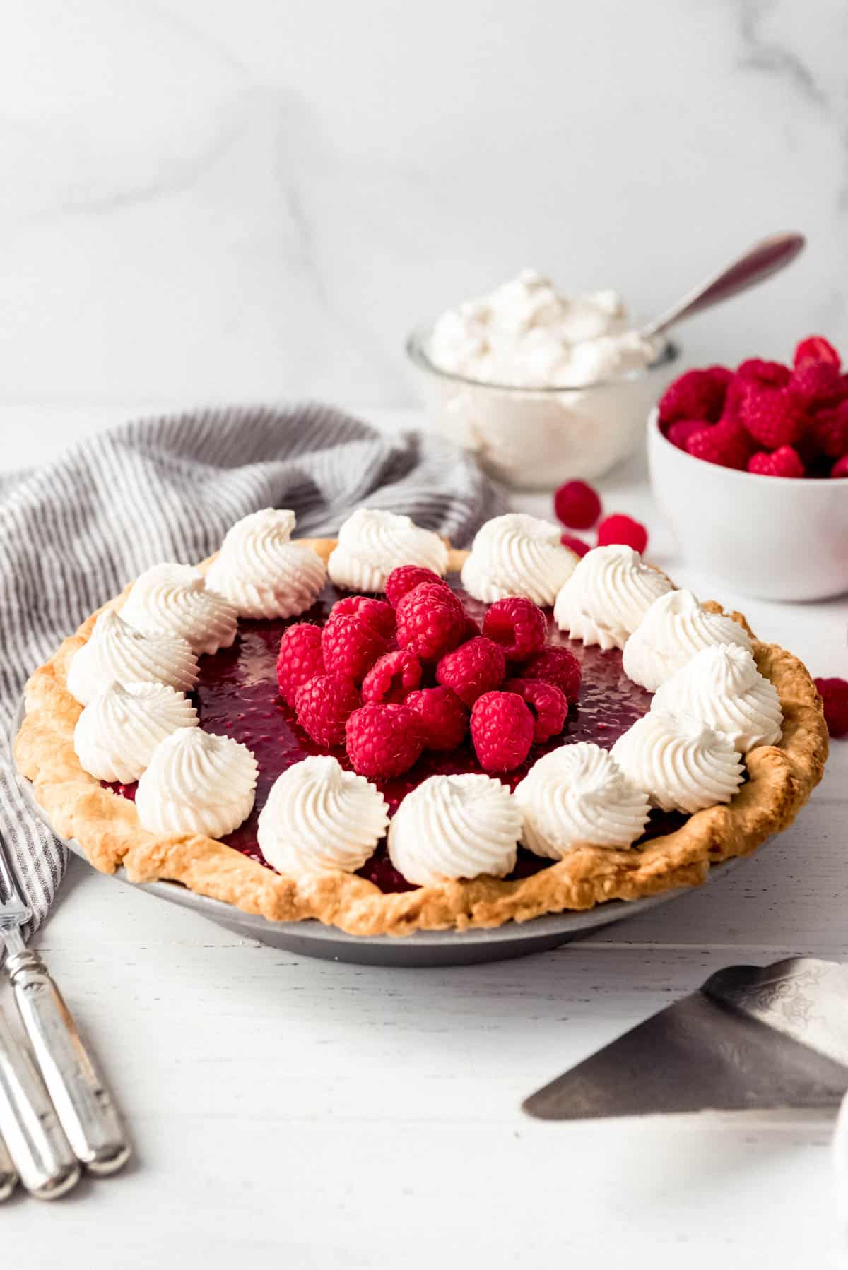 a raspberry pie topped with whipped cream in front of bowls of whipped cream and raspberries