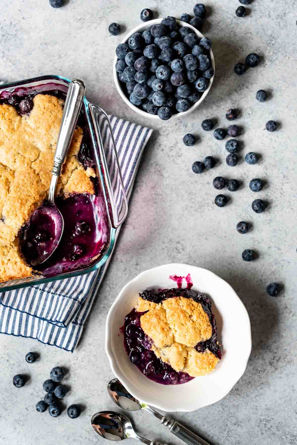 Overhead view of a dish of cooked blueberry cobbler with a scoop taken out and a silver spoon resting inside. A serving of cobbler is in a white bowl and fresh blueberries are in a separate bowl, with blueberries scattered around the dishes. 