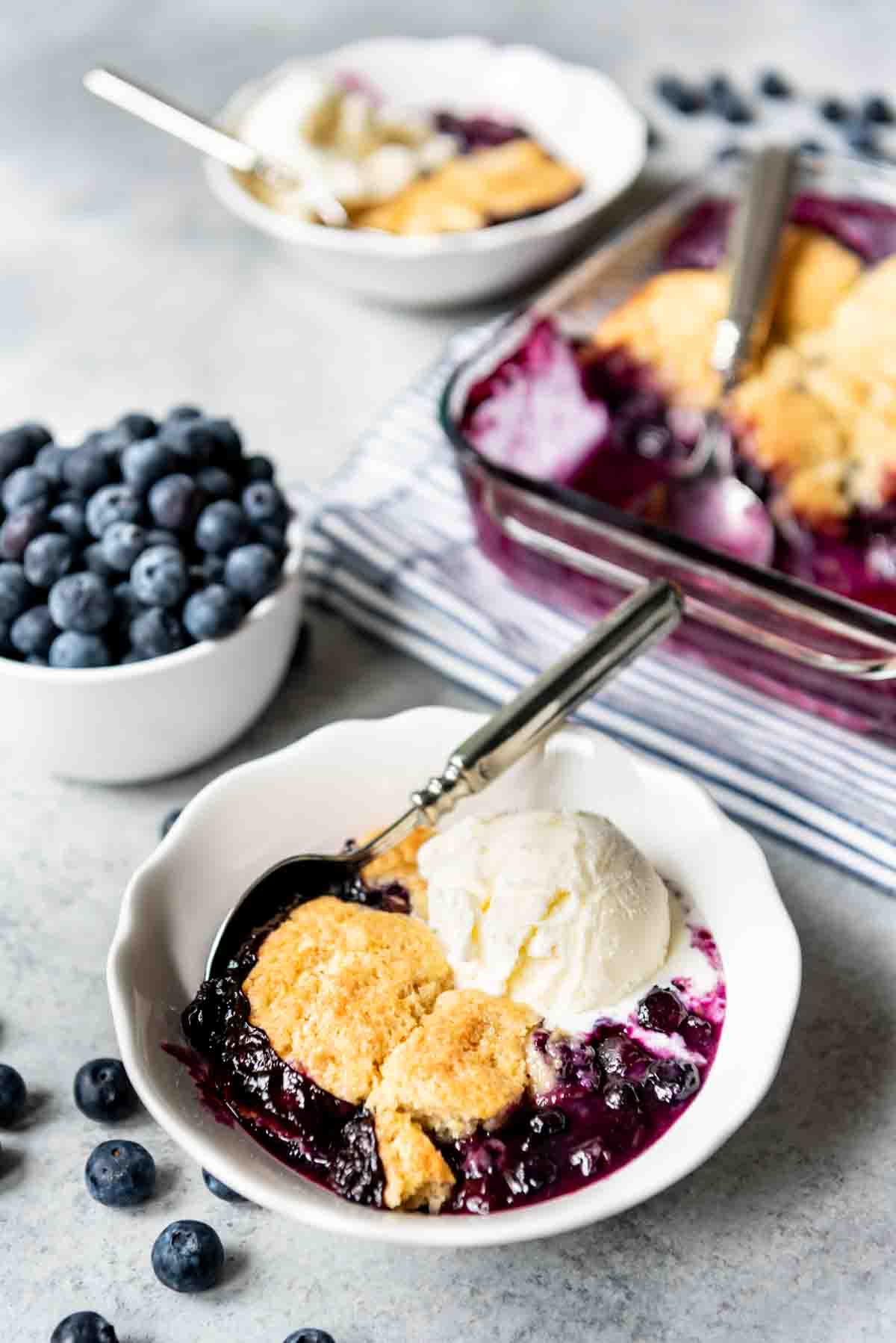 A serving of old-fashioned blueberry cobbler is placed in a bowl with a scoop of vanilla ice cream and a metal spoon. 