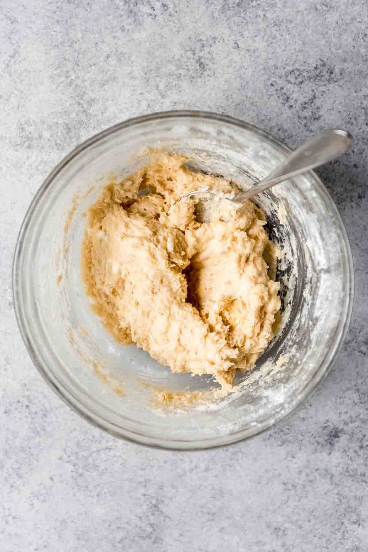 Blueberry cobbler batter is mixed and resting in a clear glass bowl, with a silver spoon inserted into the batter. 