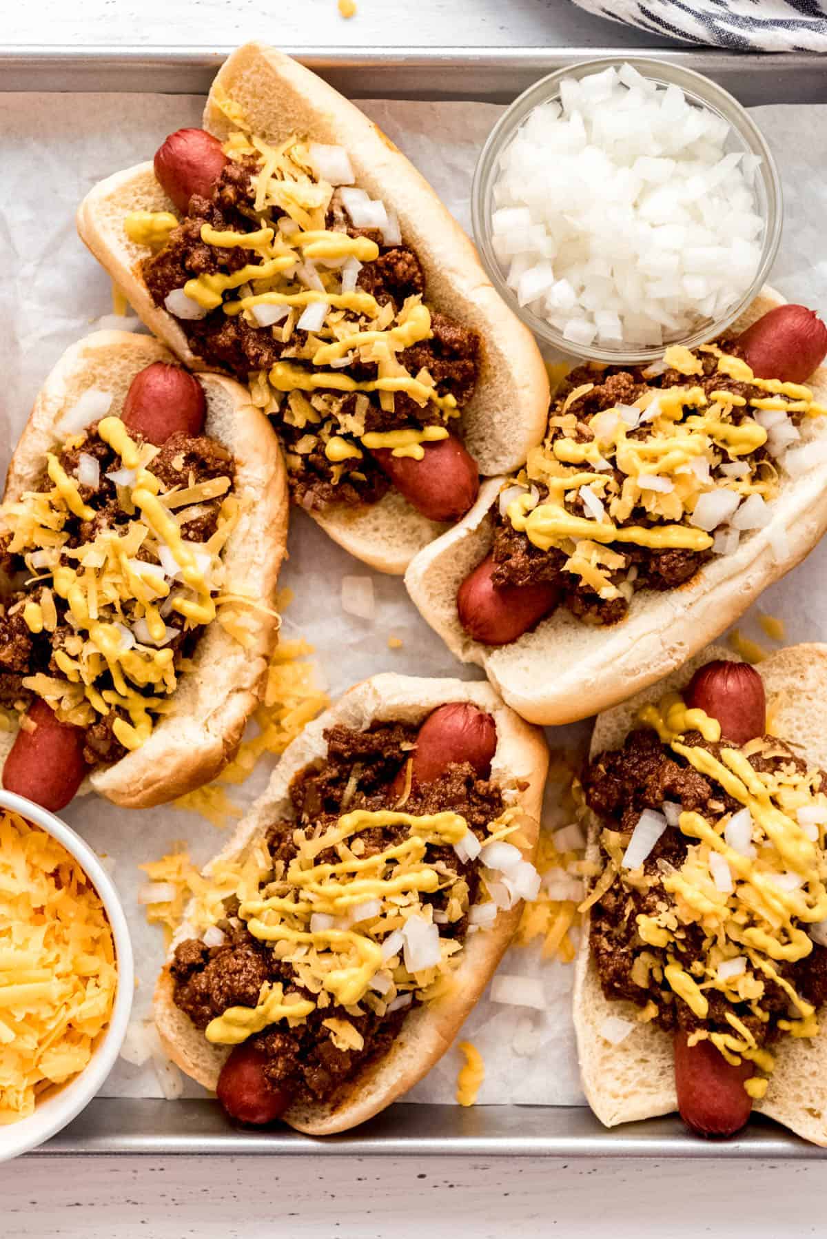 Coney Island hot dogs assembled on a baking sheet with a bowl of cheese and a bowl of onions
