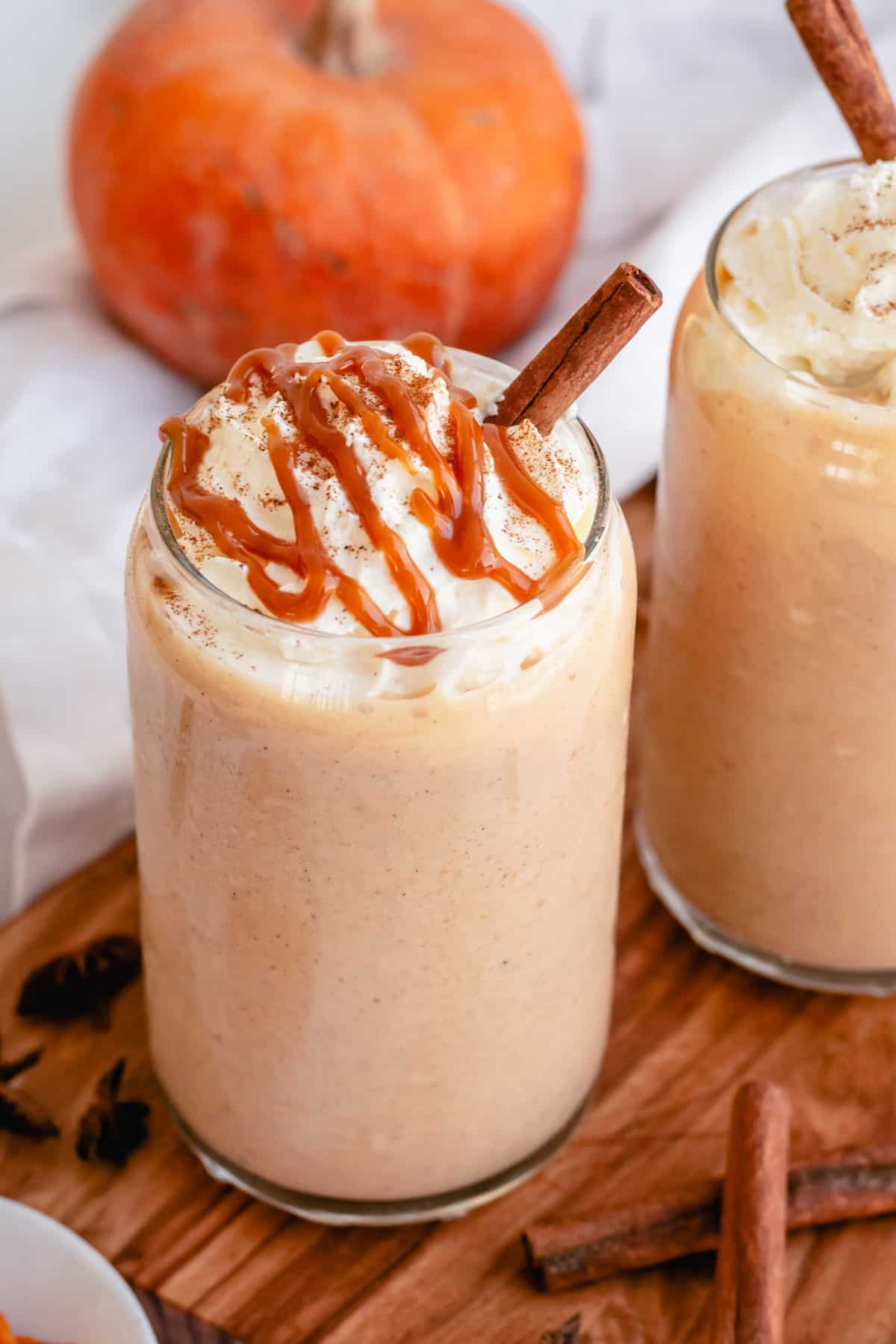 Single servings of pumpkin pie smoothies are in clear glasses, with whipped cream and caramel sauce drizzled on top. A cinnamon stick rests in the drinks, and a small pumpkin is visible in the background. 