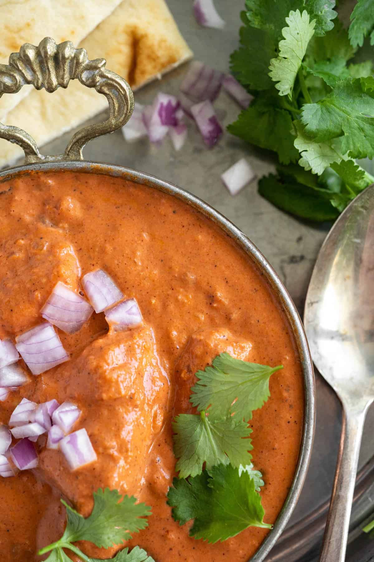 Overhead view of a bowl of Butter Chicken in a silver bowl. Diced red onion and cilantro are sprinkled on top of the dish, and a silver spoon and more cilantro are resting on the side. 