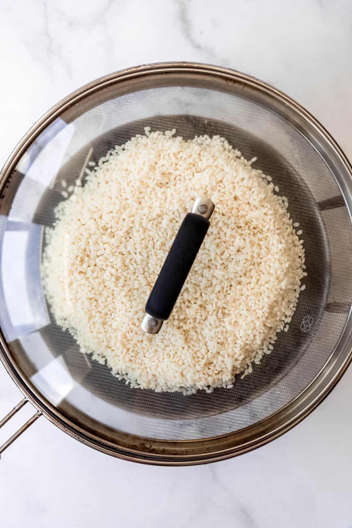 Top view of rice in a strainer sitting on top of a large pot of water with a glass lid on it. 