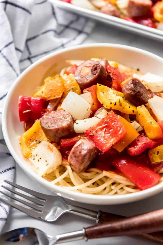 A white pasta bowl with spaghetti noodles topped with sausages and peppers next to forks..