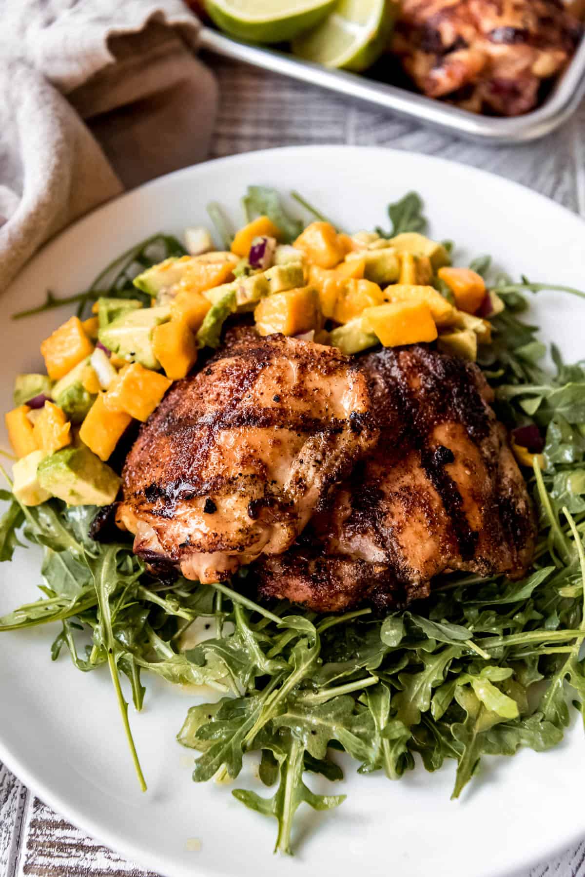 Spicy honey chicken pieces are resting on a bed of arugula salad and topped with diced mango and avocado, all resting on a white dinner plate. 