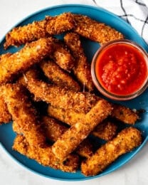 A plate full of fried mozzarella sticks next to a cup of marinara sauce.