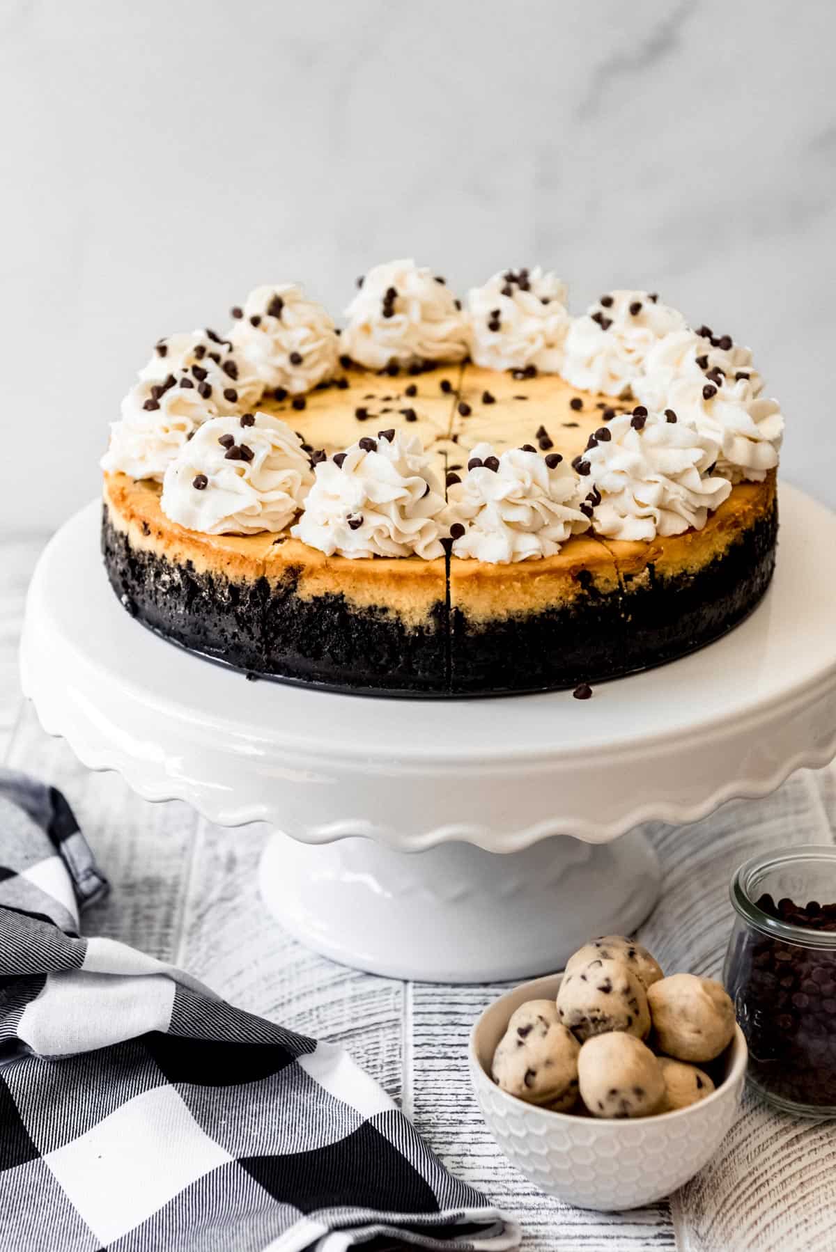 Cookie dough cheesecake on a cake stand, with balls of cookie dough in a small dish in the foreground. 