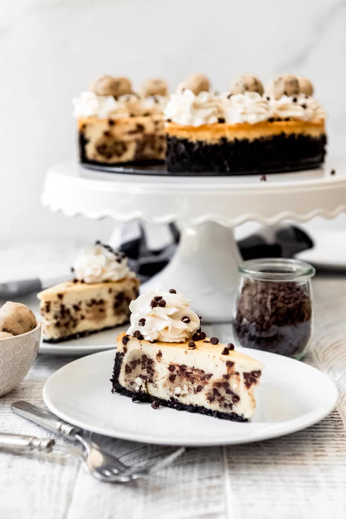 A slice of cookie dough cheesecake is on a plate, with another slice in the background and the remaining cheesecake on a cake stand. 