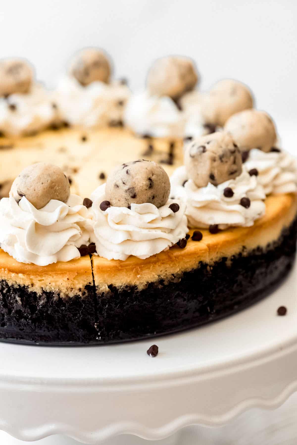 Close up view of a decorated chocolate chip cookie dough cheesecake, with whipped cream, cookie balls and mini chocolate chips on top. 