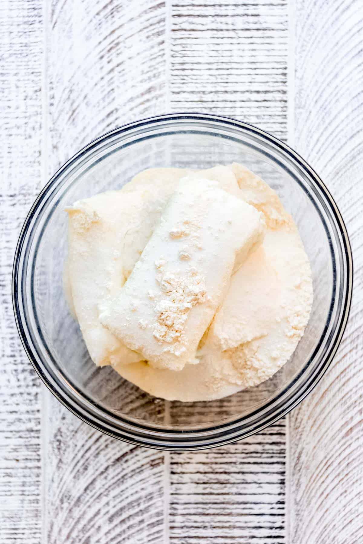 Softened cream cheese, sugar and flour are resting in a clear glass bowl, ready to be mixed. 