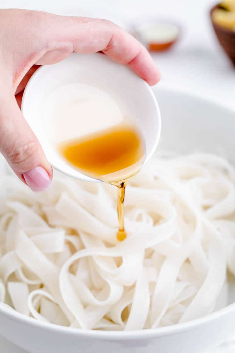 Adding sesame oil to soaked rice noodles in a bowl.