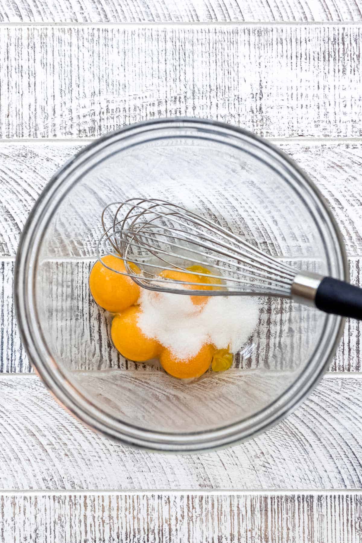 Egg yolks and granulated sugar are resting in a clear glass bowl with a wire whisk. 